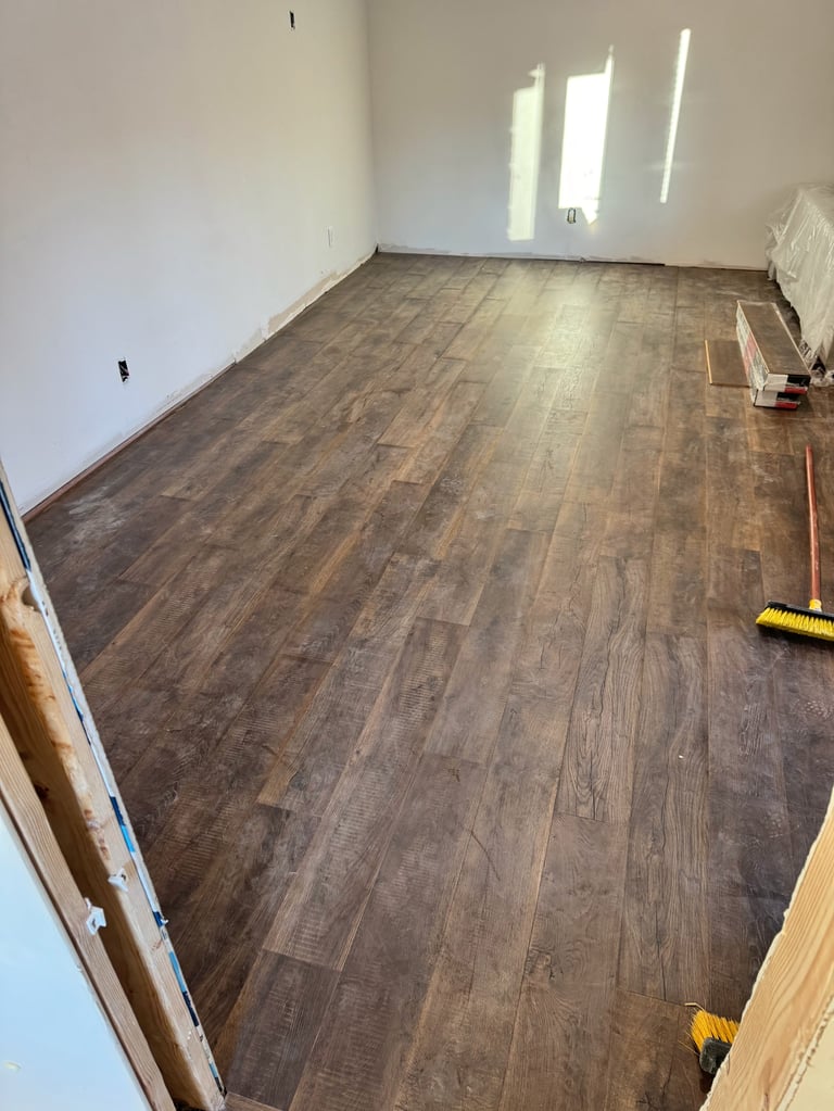Empty room with dark wood laminate flooring, white walls, and windows with natural light streaming in. Construction materials visible on the floor.