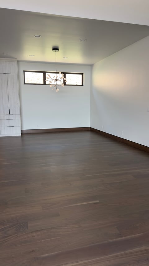 Empty modern room with dark hardwood floors, white walls, ceiling with recessed lighting, and high window with decorative pendant light