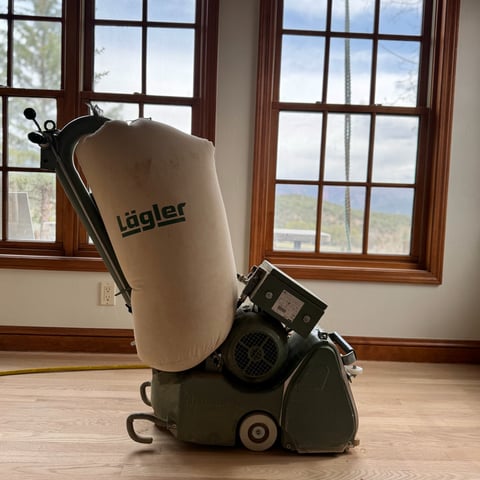 Lagler hardwood floor sanding machine with cream dust collection bag positioned in front of wooden windows in an interior room.