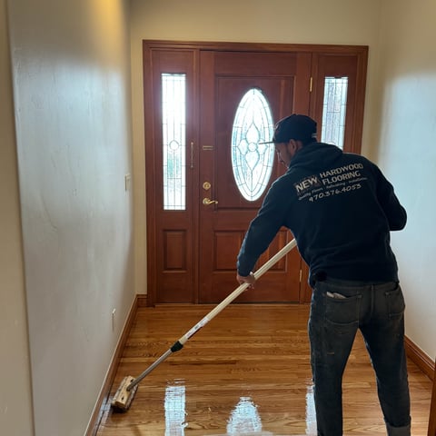 Worker refinishing hardwood floor in entryway using floor sander, with wooden door with circular window in background
