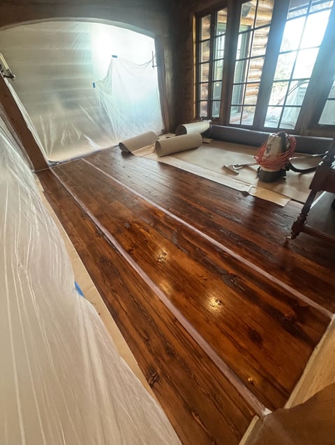 Empty room with dark hardwood floors, plastic sheeting, rolled materials, and large windows with panes showing renovation in progress