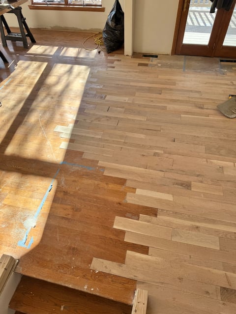 Indoor room during hardwood floor installation with partially installed wood planks, sunlight streaming through windows, and construction materials visible.