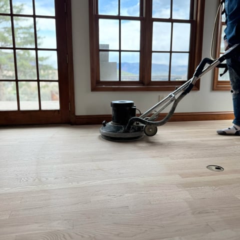 Worker operating a floor buffer machine on wooden flooring near large windows with water view